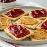 Peanut butter and jelly spread on rectangular crackers arranged on a ceramic plate.