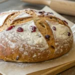 A rustic round loaf with a golden crust, lightly dusted with flour and studded with red berries, resting on parchment paper atop a wooden cutting board.
