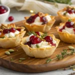 Mini phyllo cups filled with goat cheese and cranberries, garnished with herbs and nuts, arranged on a wooden board.