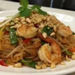 Thai shrimp stir-fried noodles garnished with peanuts, vegetables, and fresh cilantro on a white plate.